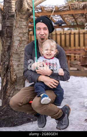 Kanadischer Vater lächelt der Kamera zu, während er auf einer Schaukel spielt, während sein Kleinkind auf seinem Schoß sitzt und ohne Grund weint, in einem verschneiten Spätwintergarten Stockfoto