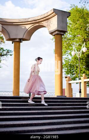 Eine dünne weibliche Ballerina in einem rosa Seidenkleid und spitzenschuhe mit Bändern senkt sich anmutig die Stufen in einem Bogen mit Säulen im Park hinunter Stockfoto