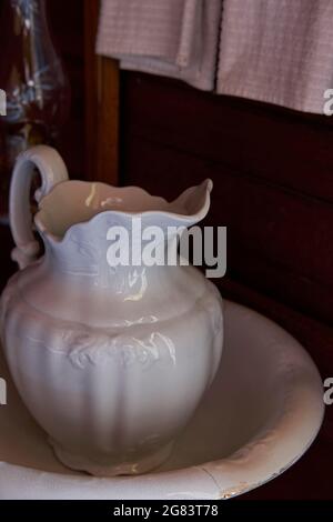 Ein weißes antikes Porzellanwaschbecken und ein Krug ruhen auf einem hölzernen Waschtisch. Weiße Handtücher hängen im Hintergrund. Stockfoto
