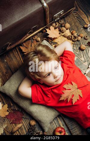 Kind hat Spaß mit Leaf Fall. Netter Junge, der im Herbst spielt. Das Kind liegt mit den Händen hinter dem Kopf und ruht sich in Blättern auf dem Holzboden aus. Herbstkonzept Stockfoto