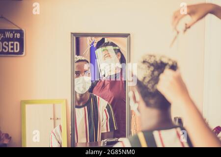 afrikanischer Mann beim Friseur, der wegen einer Coronavirus-Pandemie eine Schutzmaske trägt. Junge asiatische Frau Haare schneiden in covid Zeit. Stockfoto