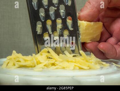 Nahaufnahme der Hand eines Mannes, der ein Stück Cheddar-Käse auf ein rundes Marmorbrett reiben soll. Stockfoto