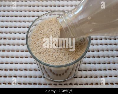 Nahaufnahme eines schaumigen, milchigen, vereisten Kaffee-Latte, der aus einer Plastikflasche aus dem Kühlschrank in ein Trinkglas gegossen wurde. Stockfoto