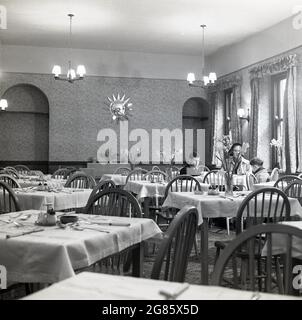 1950s, historisch, eine Dame und ihre zwei kleinen Kinder, die an einem Tisch im oberen Stockwerk eines Cafés oder Teestuben der Zeit mit mit Stoffgedecken bedeckten Tischen und Narzissen in Glasblumentöpfen, England, saßen. VEREINIGTES KÖNIGREICH. Stockfoto