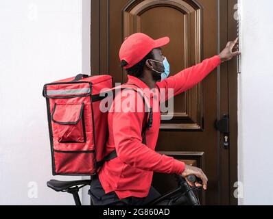 Afrikanischer Fahrer, der beim Ausbruch des Corona-Virus eine Mahlzeit mit einem Elektrofahrrad an die Kunden nach Hause liefert, während er eine Gesichtsmaske trägt Stockfoto