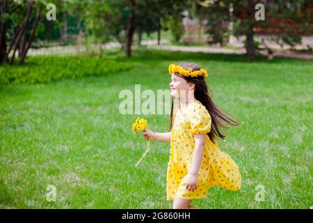 Ein lächelndes Mädchen in einem gelben Kleid und gelben Blumen mit geschlossenen Augen genießt einen warmen, sonnigen Tag im Hinterhof eines Dorfhauses Stockfoto