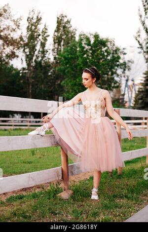 Eine junge, elegante Ballerina setzt ihren Fuß auf einen Holzzaun und bindet an einem warmen Tag Bänder auf spitzenschuhe im Park Stockfoto