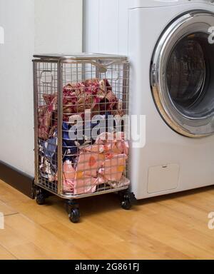 Wäschekorb aus Edelstahl mit Rädern und schmutzigen Tüchern in der Nähe der Waschmaschine im Waschraum Stockfoto