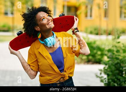 Multiethnische Frau mit lockigen Haaren hält Skateboard während des Spazierens moderne Stadt genießen Aktivität Freizeit Sommer Stockfoto