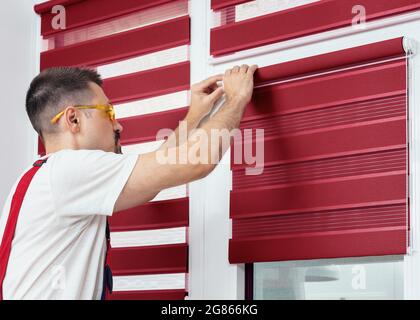 Montage von Rollläden, Mann, der Stoffjalousien installiert. Handwerker, der einen Teil des Rollfensters in Innenräumen installiert. Bauarbeiter Mann in einer Uniform Stockfoto