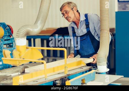 Ein älterer Tischler arbeitet in einer Schreinerei. Sägen von Holzbrettern. Authentische Szene Rentner in Brille bei der Arbeit. Stockfoto