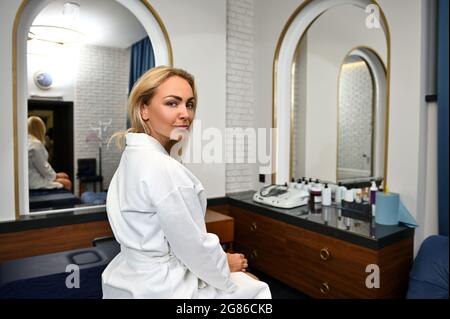 Entzückende junge europäische Frau in weißem Gewand, die über die Schulter auf die Kamera blickt, während sie sich im Schönheitssalon entspannt Stockfoto