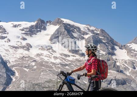 Nette und aktive ältere Frau, die auf dem Pralongia Plateau in den Dolomiten mit dem Gletscher des Marmolata-Gipfels in Backgrou ihr elektrisches Mountainbike fährt Stockfoto