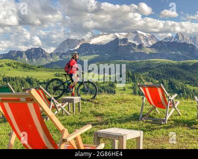 Nette und aktive ältere Frau, die auf dem Pralongia Plateau in den Dolomiten mit dem Gletscher des Marmolata-Gipfels in Backgrou ihr elektrisches Mountainbike fährt Stockfoto