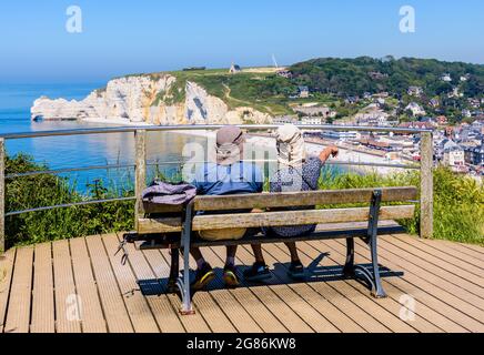 Ein paar Wanderer sitzen auf einer Bank und genießen an einem sonnigen Tag den Panoramablick über die Strandstadt Etretat und die Klippe Amont in der Normandie. Stockfoto