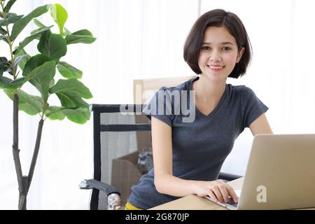 Junge, hübsche und wunderschöne Thai-deutsche Halbrennerin, die als Schreibtisch sitzt und lächelt, während sie im Schlafzimmer zu Hause einen Labtop-Computer benutzt. Idee für Zufall Stockfoto