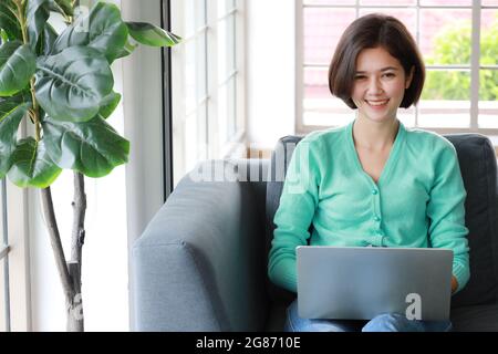 Junge, hübsche und wunderschöne Thai-deutsche Halbrennerin, die als Schreibtisch sitzt und lächelt, während sie im Wohnzimmer mit einem Labtop-Computer auf dem Sofa arbeitet. Idee für ha Stockfoto