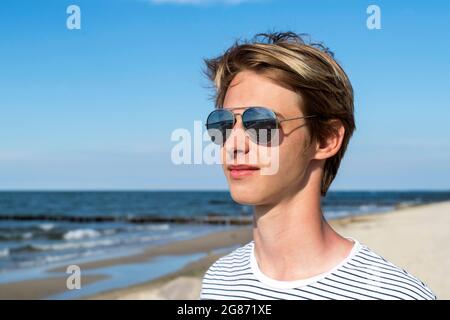 Blonde Teenager-Junge in Spiegelbrille auf dem Sandstrand stehen Stockfoto