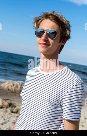 Blonde Teenager-Junge in Spiegelbrille auf dem Sandstrand stehen Stockfoto