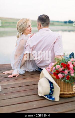 Rückansicht eines gutaussehenden Mannes mittleren Alters und einer hübschen, charmanten blonden Dame, die auf der Holzbrücke am Ufer des Sees oder des Teichs sitzen und sich mit Liebe anschauen. Stockfoto