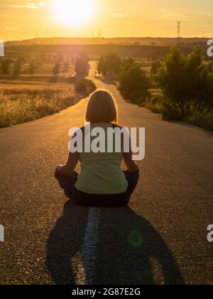 Vertikale Aufnahme einer jungen Frau, die mit ihrem Rücken auf einer Straße sitzt und dem Sonnenuntergang gegenübersteht Stockfoto
