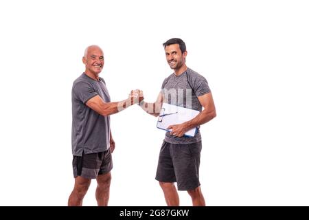 Fitnesstrainer mit Workout-Pad schüttelt sich die Hände mit einem pensionierten Mann in Sportuniform. Auf einem weißen, isolierten Hintergrund. Stockfoto