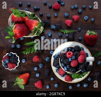 Beerenmischung auf einem hölzernen Hintergrund. Beeren in einer Tasse und Schüssel. Erdbeeren, Blaubeeren, Brombeeren und Himbeeren. Sommerhintergrund. Draufsicht, FLA Stockfoto