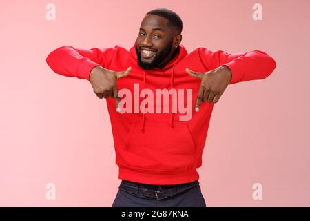 Freche junge charismatische gut aussehende bärtige schwarze Kerl in roten Hoodie Zeigen nach unten Zeigefinger lächeln breit zeigt genial Kopieplatz Die Lösung ist cool Stockfoto