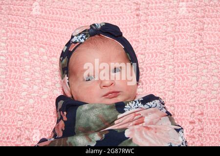 Nahaufnahme Porträt eines neugeborenen Mädchens mit braunen Haaren trägt geblümten Stirnband in Decke des gleichen Musters gewickelt Blick auf den Betrachter. Auf einem liegen Stockfoto