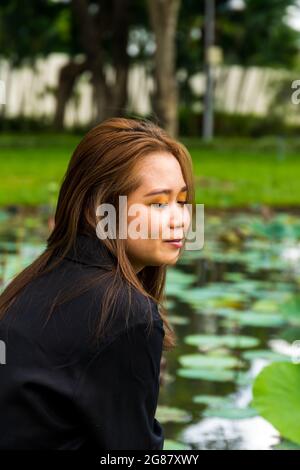 Ein Porträt einer jungen südasiatischen Frau, die neben einem Teich mit Seerosen im Park posiert Stockfoto