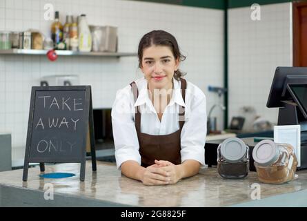 Die jungen kaukasischen Mitarbeiter im Café mit brauner Schürze lehnen sich an die Theke, während sie auf die Kunden warten, die den Laden betreten. Morgenstimmung in einem Stockfoto