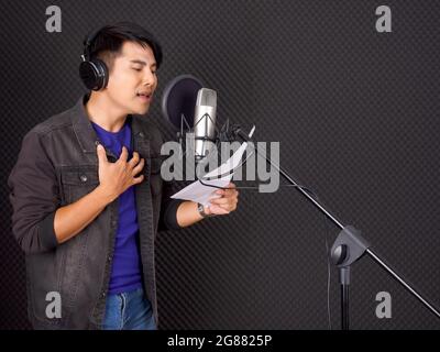 Musiker, die Musik im professionellen Aufnahmestudio produzieren. Ein junger asiatischer Mann mit Kopfhörern singt, während er den Text in der Hand vor dem schwarzen so hält Stockfoto