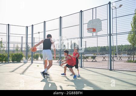 Drei Brüder spielen Basketball, einer von ihnen hat eine Beinprothese. Geschwister spielen Korb. Stockfoto