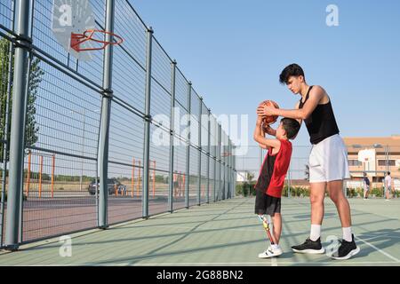 Basketballtrainer zeigt einem Kind mit einer Beinprothese, wie man Basketball schießt. Coach, der ein Kind trainiert. Stockfoto