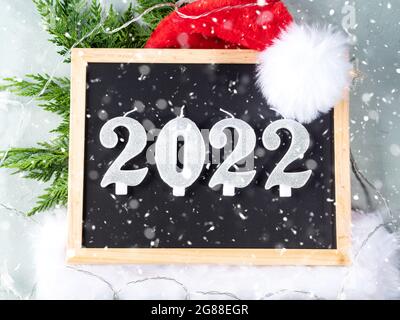 Neujahr 2022 Zahlen auf Tafel mit weihnachtsmütze und Tannenzweigen auf grau mit Sternenlicht Stockfoto