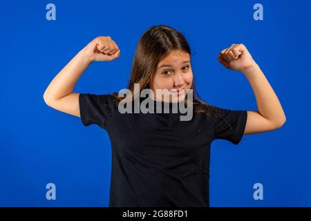 Hübsches Mädchen, das ein schwarzes T-Shirt trägt und dabei Muskeln auslegt, um das stärkste Mädchen der Welt zu sein, isoliert auf blauem Studiohintergrund Stockfoto