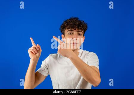 Junger Mann mit lockigen Haaren, der auf eine Ecke des Bildes zeigt, in der der Text des Kopierraums auf blauen Studiohintergrund geht. Marketingkonzept Stockfoto