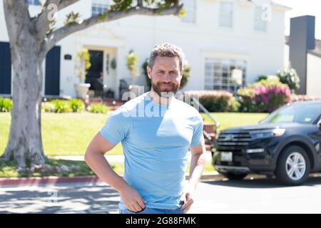 Lächelnder bärtiger Mann im lässigen blauen Hemd draußen bei Haus und Auto, gute Laune Stockfoto