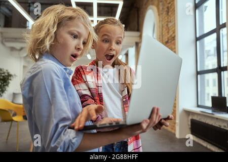 Zwei reizende Kinder, ein kleiner Junge und ein Mädchen, die aufgeregt mit einem Laptop unterwegs sind und während des MINT-Unterrichts in einem Klassenzimmer stehen Stockfoto