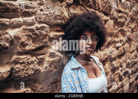 afro-Frau mit Brille und lässiger Kleidung, die die Kamera anschaut. Sie lehnt sich an eine Steinmauer Stockfoto