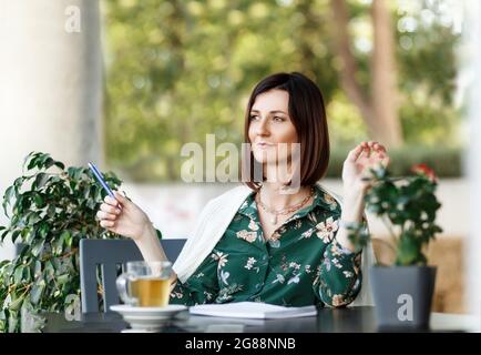 Eine junge, selbstbewusste Frau sitzt mit einer Tasse grünem Tee am Tisch. Stadtcafé. Das Mädchen verbringt ihre Freizeit in einer gemütlichen Atmosphäre. Ein durchdachtes Aussehen. Stockfoto