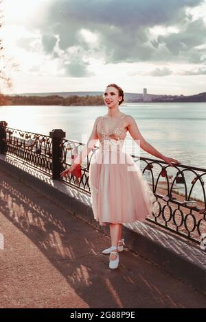 Eine lachende, lächelnde Ballerina in einem rosa Seidenkleid hält sich an einem sonnigen Sommertag am Zaun des Pfades entlang der Wolga bei Sonnenuntergang fest Stockfoto