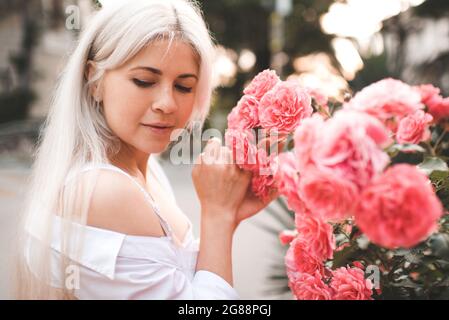 Schöne blonde Frau 29-30 Jahre alt mit langen blonden Haaren riechen Blumenrosen im Garten im Hintergrund aus nächster Nähe. Sommerferiensaison. Stockfoto