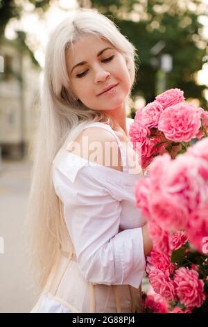 Attraktive blonde Frau 25 - 29 Jahre alt hält rosa Rosenblüten im Park im Freien. Gesunde, natürliche Haut. Frühjahrssaison. 20s. Urlaubszeit. Stockfoto