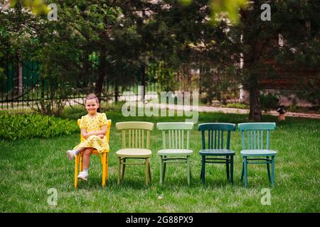 Ein lächelndes Mädchen in einem gelben Kleid sitzt mit gekreuzten Beinen auf bunten Wiener Stühlen und steht in einer Schlange im Kinderhausentheater und wartet auf ein Theaterstück Stockfoto