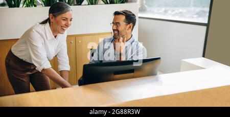 Glücklicher Manager, der sich mit einem Kollegen an der Rezeption unterhielt. Reife Frau hilft jungen Mann bei der Arbeit am Computer am Schreibtisch. Stockfoto