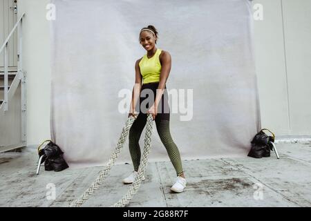 Afrikanische Frau in Fitness Tragen Sie Crosstraining mit Kampfseil. Weibliche Athletin macht Kampfseil Workout-Übung, Blick auf die Kamera und lächeln. Stockfoto