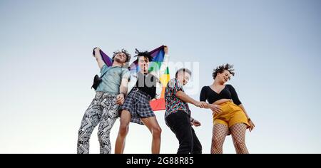 Fröhliche Gruppe von queeren Individuen, die den Stolz der Homosexuellen feiern. Vier Mitglieder der LGBTQ+ Community tanzen fröhlich und heben die Regenbogenflagge. Stockfoto