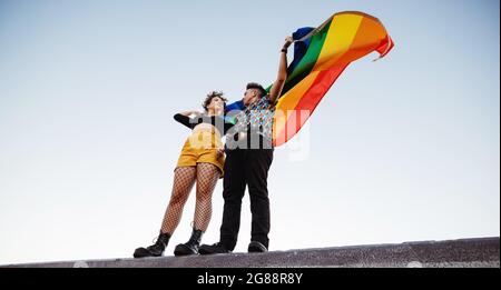 LGBTQ-Paar, das draußen die Regenbogenfahne hochhebt. Queere junge Männer und Frauen, die ihre LGBTQ+-Identitäten annehmen. Junge nicht konforme Paar steht w Stockfoto