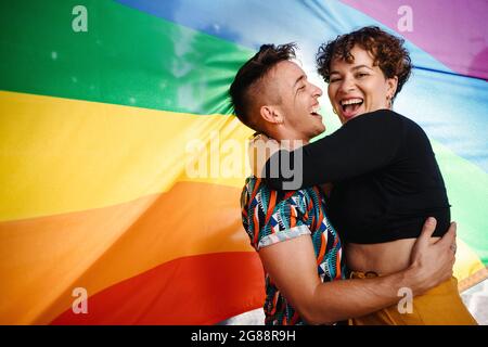 Fröhliches, queeres Paar, das gegen eine Regenbogenflagge steht. Das junge LGBTQ-Paar lächelt fröhlich, während es sich umarmt. Zwei nicht konforme Liebhaber Stockfoto